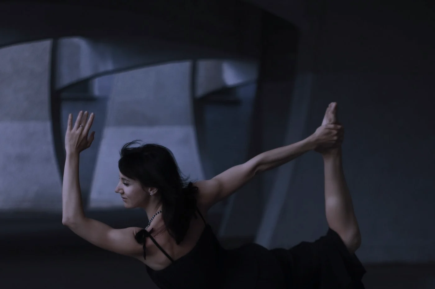 A woman mid-movement, one arm and one leg extended, against concrete in low light.